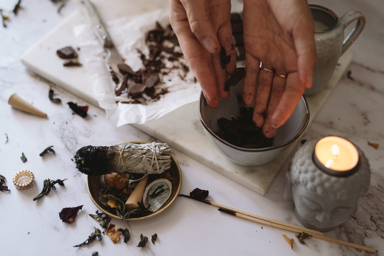 Hands preparing herbs for aromatherapy beside burning sage and candle on marble surface.