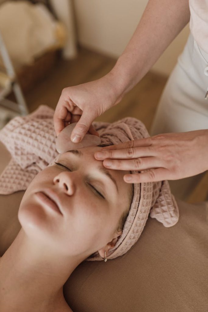 woman-lying-with-a-towel-on-her-head-6663571 Woman enjoying a soothing facial massage with gua sha tool, promoting relaxation and skin care.